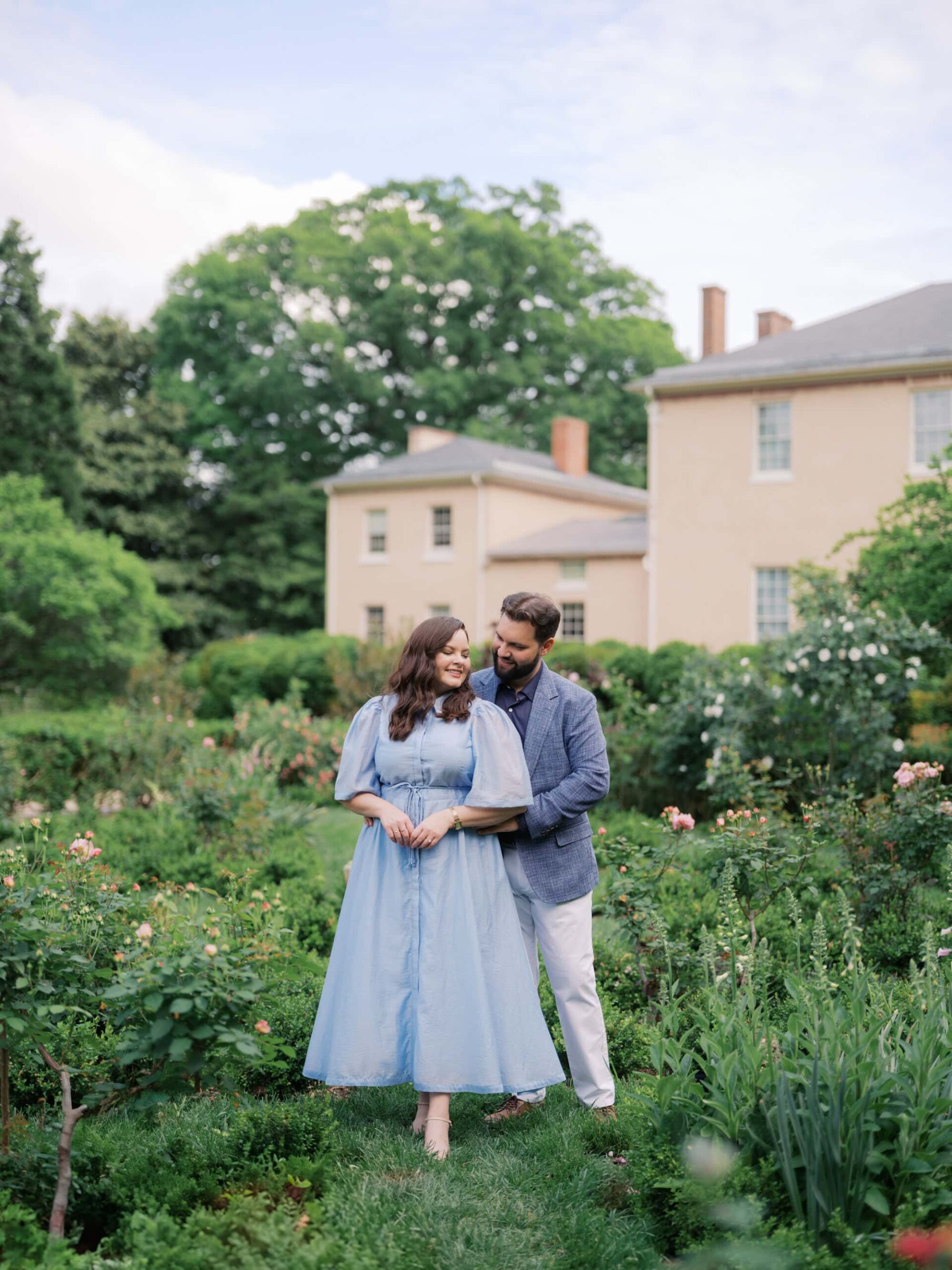 Couple posing at Tudor Place, a historic home and garden in Georgetown, one of the best engagement photo locations in DC. The woman is wearing a blue dress and the man is wearing white chinos, brown shoes and a blue jacket.