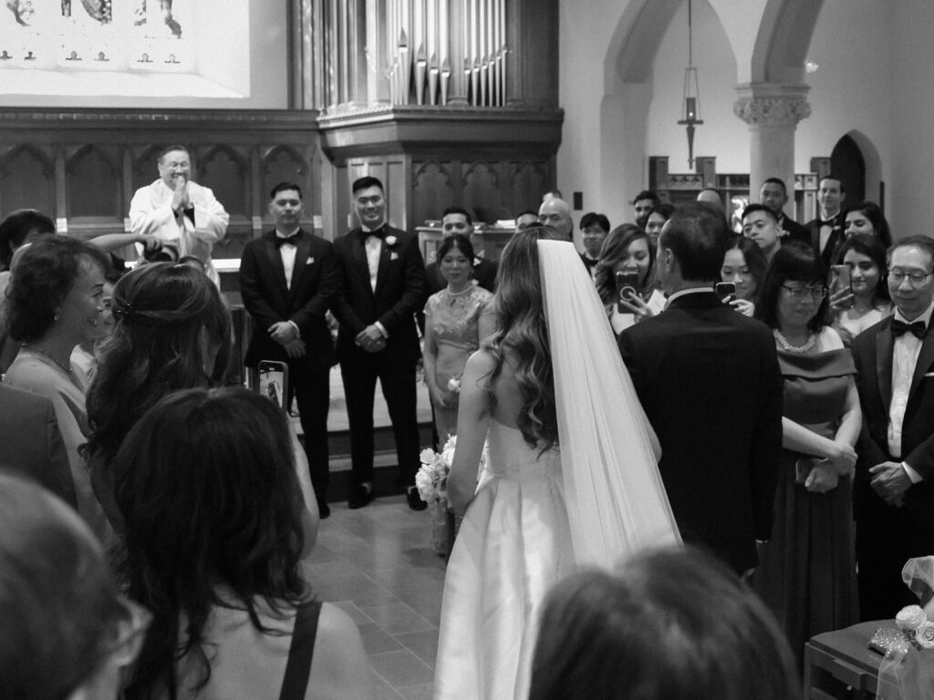 A black and white photo of a wedding ceremony, featuring the bride in a white gown and veil standing with the groom, surrounded by guests and a minister, set in a beautiful church interior.