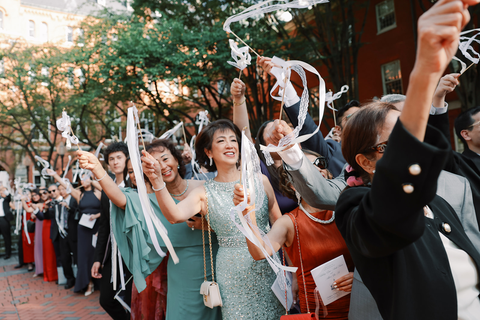 Guests celebrate with ribbon wands outside, smiling and waving them during a wedding send-off, with a backdrop of trees and brick pavement.