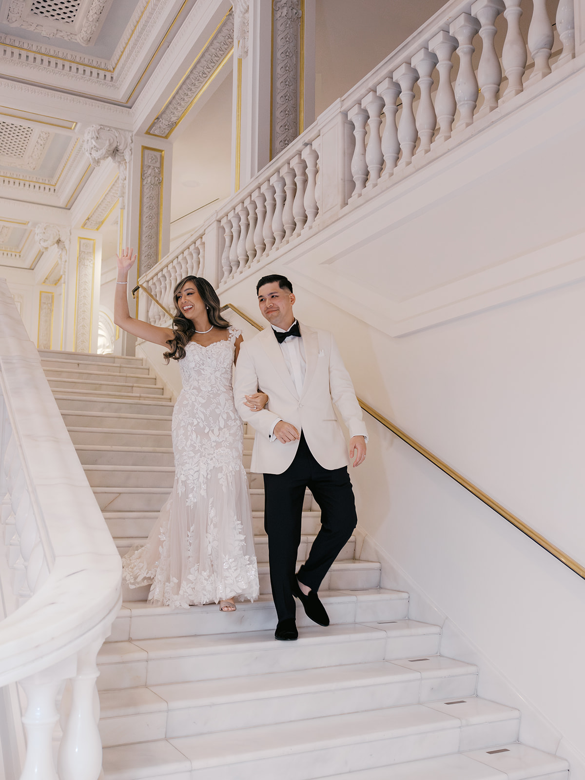 A bride in an elegant lace wedding gown and a groom in a white tuxedo walking hand in hand down a grand staircase, smiling and waving.