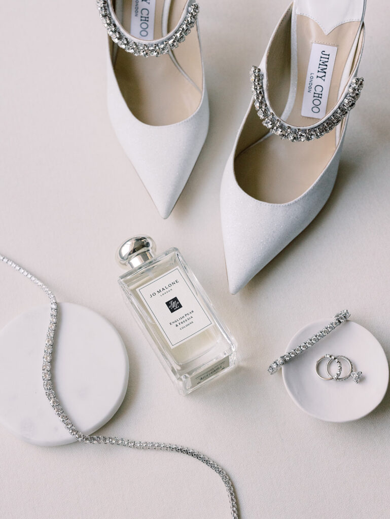 A flat lay of elegant wedding accessories including Jimmy Choo white heels, Jo Malone perfume, sparkling jewelry, and a decorative marble dish.