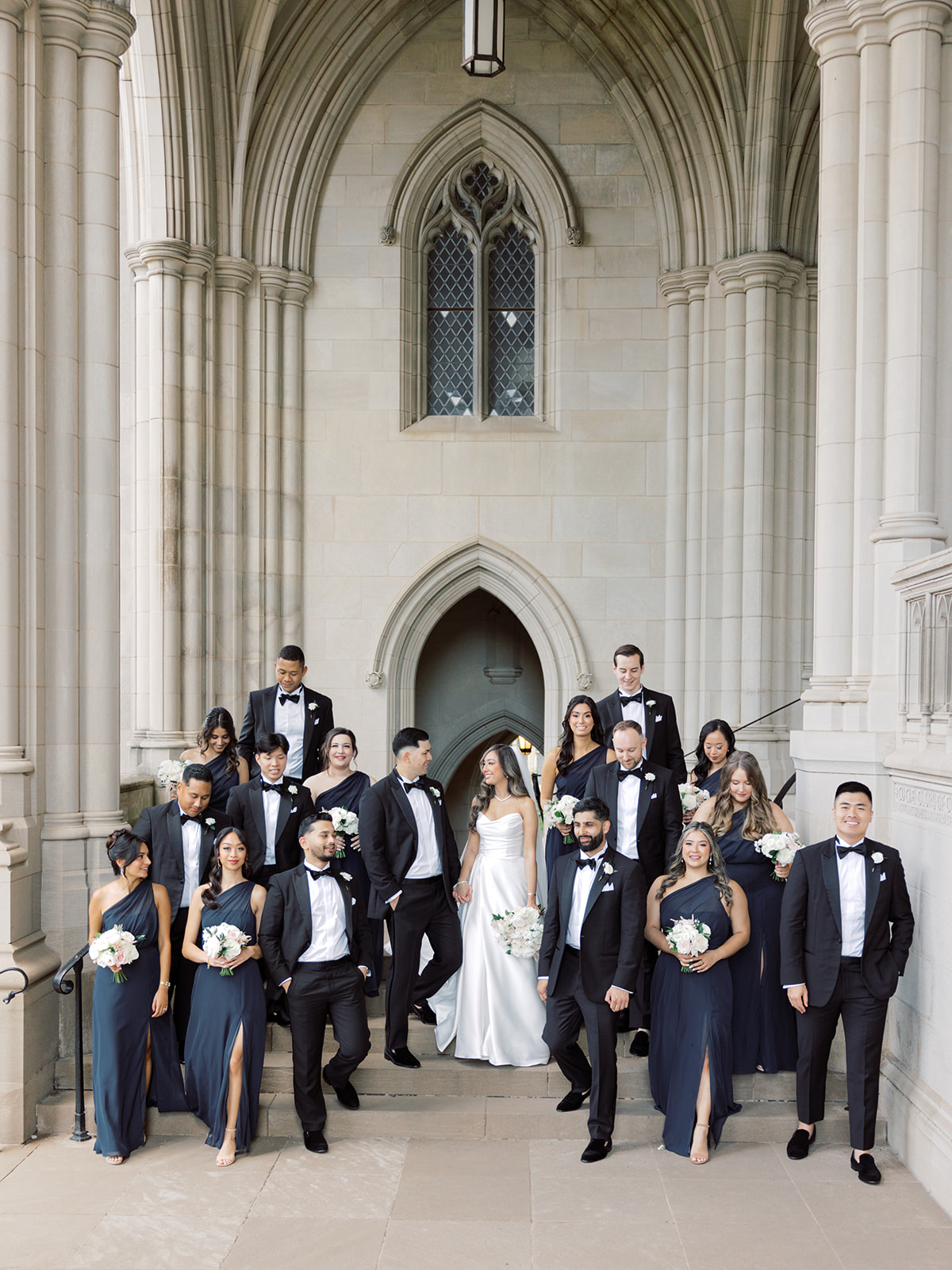 A wedding party posing on the steps of a the Washington National Cathedral, featuring a bride in a white gown and bridesmaids in navy dresses, alongside groomsmen in tuxedos.