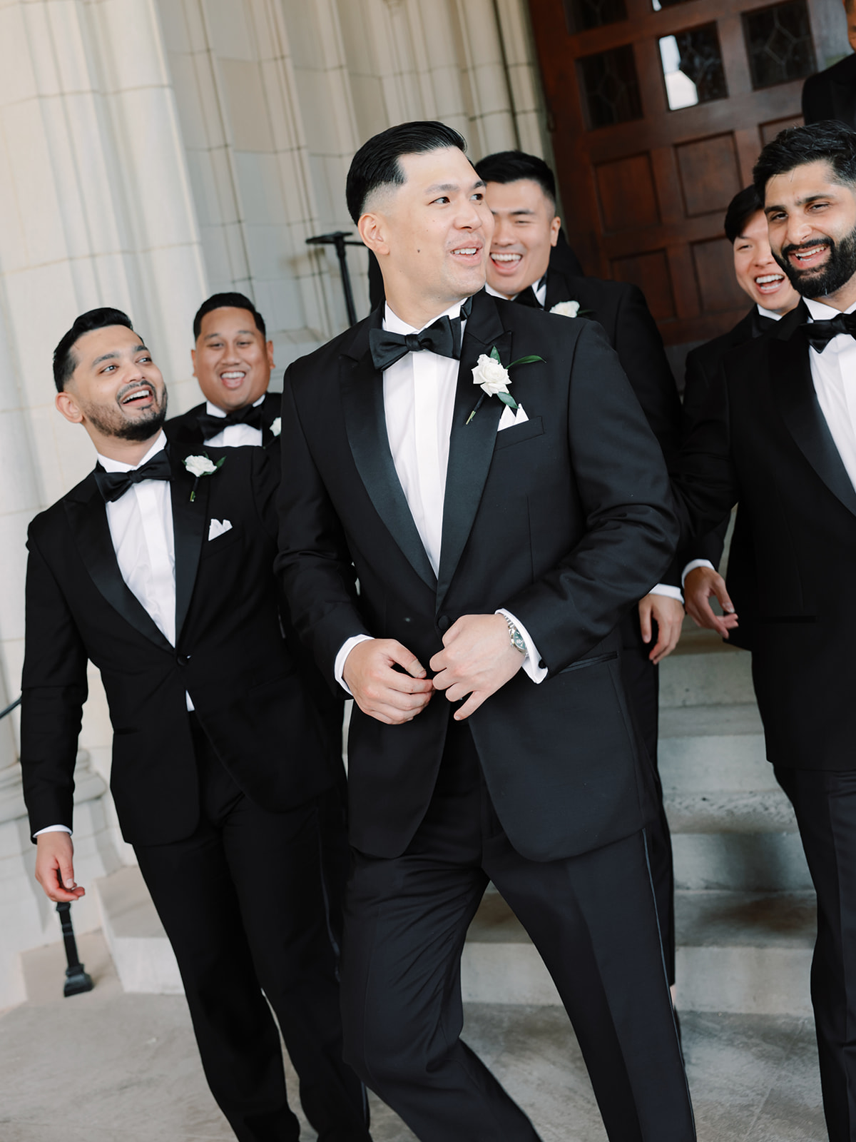 Groom smiling and walking with groomsmen in tuxedos at a wedding, standing on steps of a historic building.
