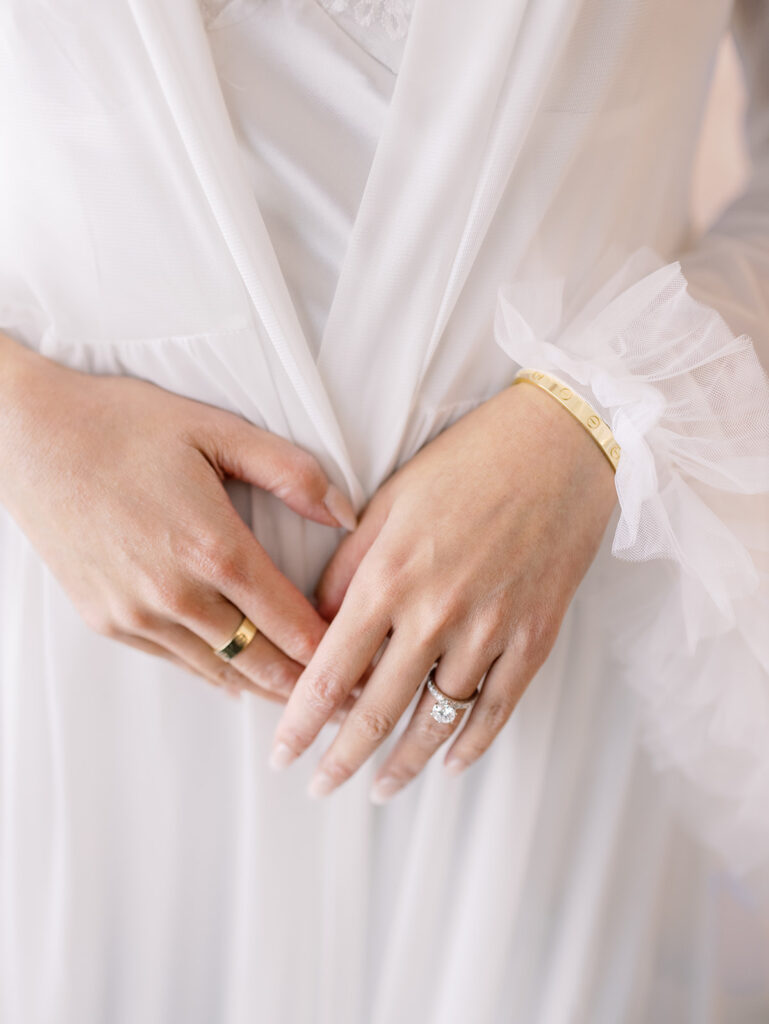 Close-up of a bride's hands wearing a gold Cartier band and an engagement ring with a large center stone, dressed in a white robe.