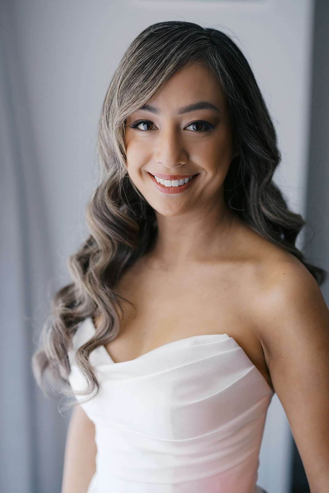 Close-up portrait of a bride with long, wavy hair wearing a strapless wedding dress, smiling brightly against a softly lit background.