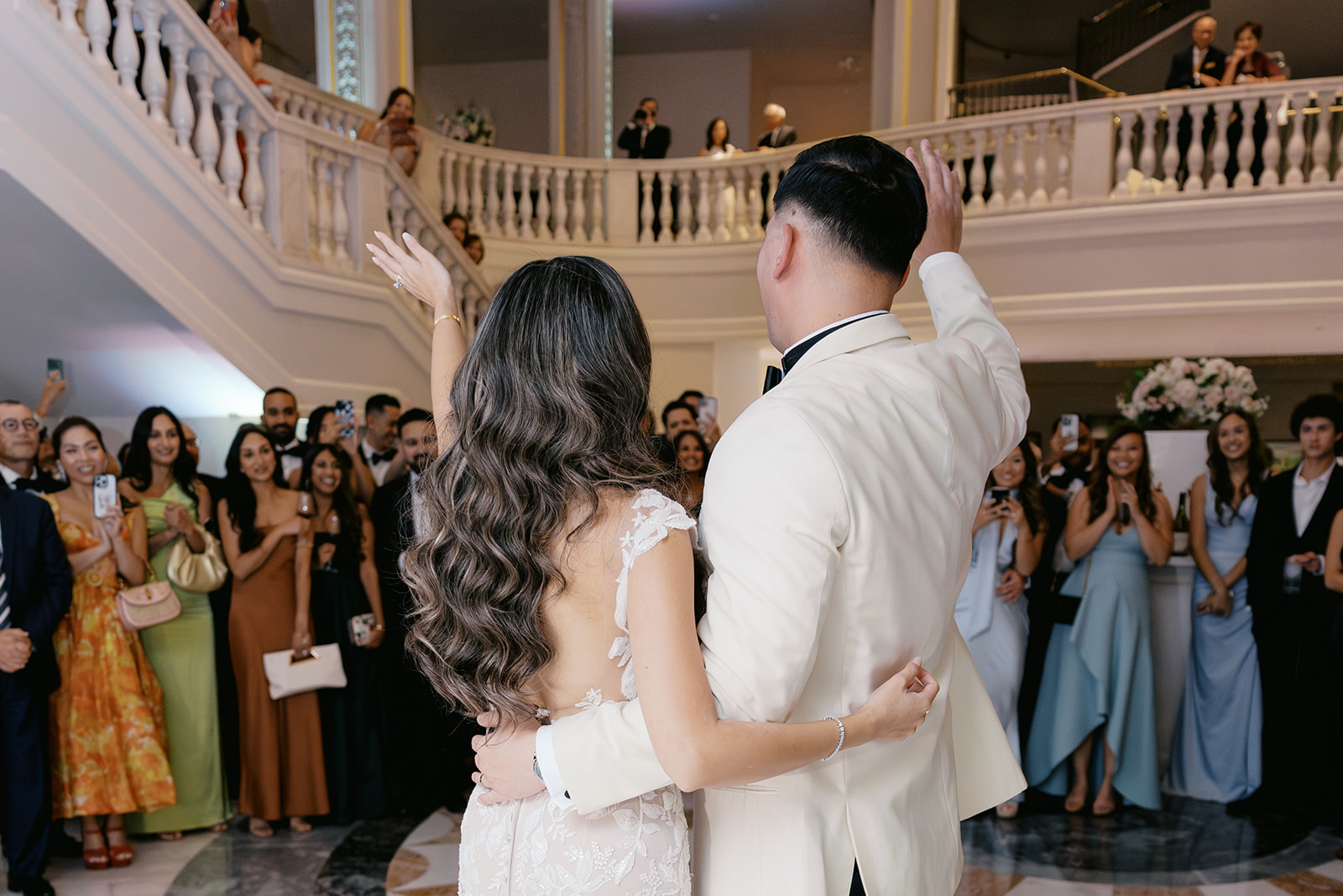 A couple stands together at their wedding reception at the National Museum of Women in the Arts (NMWA), smiling and waving to guests while they gather on the stairs, capturing the joyful atmosphere of the celebration.
