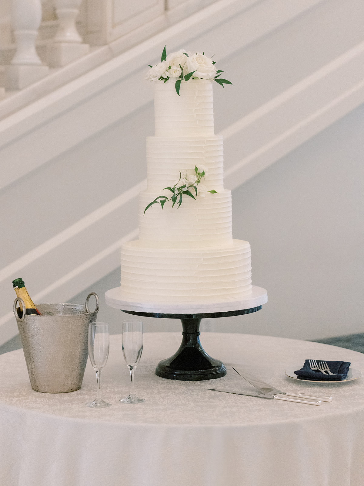 Three-tiered white wedding cake decorated with white flowers and greenery on a round table, accompanied by a champagne bucket and fluted glasses.