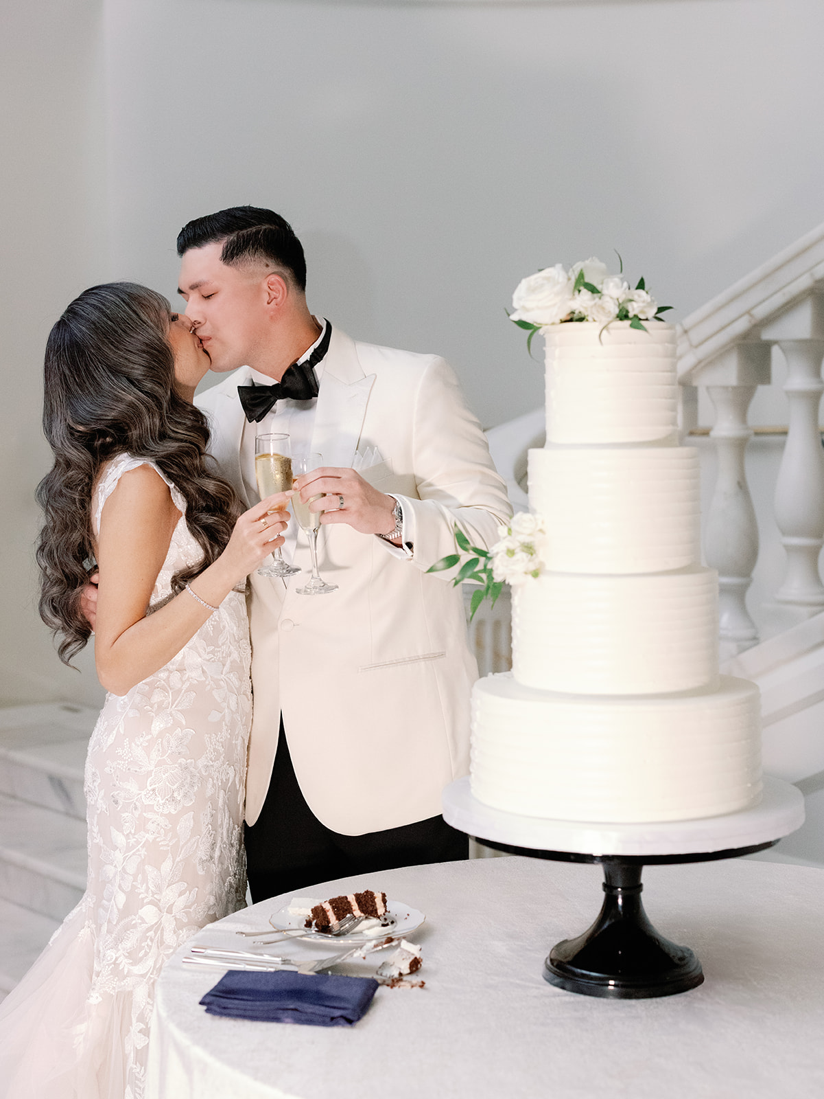 Couple sharing a kiss at their Washington DC wedding at the National Museum of Women in the Arts, while holding champagne glasses, standing next to a beautiful wedding cake adorned with white roses.